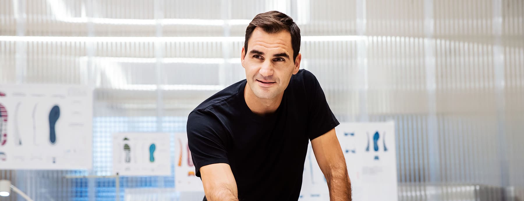 Roger Federer in a black t-shirt posing in an indoor On setting with blurred background
