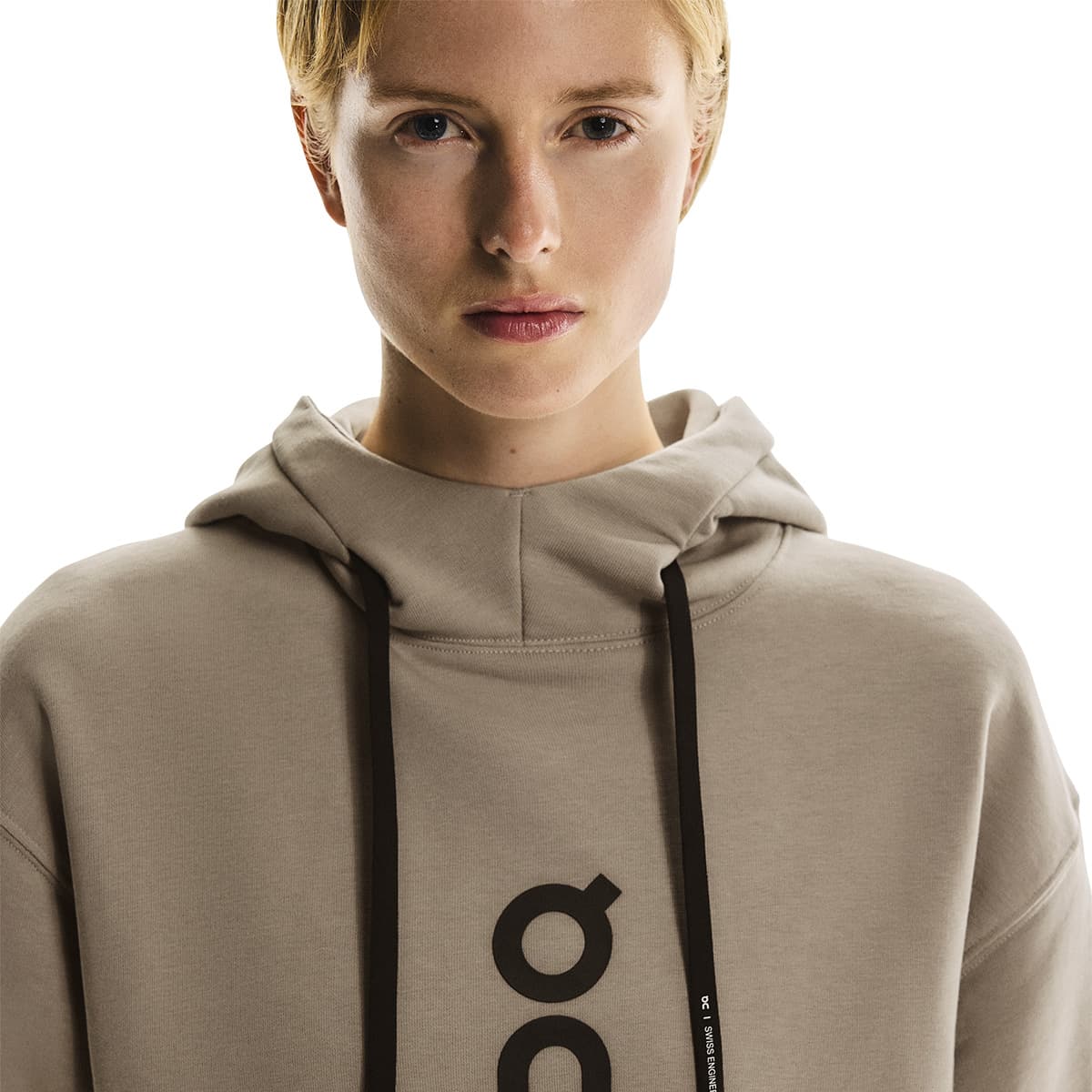 Front close-up of female model wearing the On Club Hoodie in Desert showing logo and drawstring detail